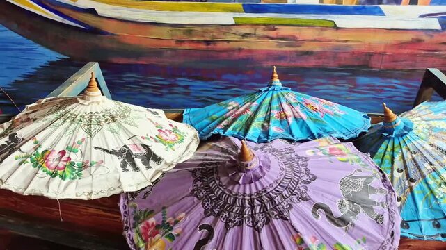 Colorful Asian paper parasols with floral and elephant motifs arranged in wooden boat against painted backdrop, cultural decorative items, traditional craft display, exotic aesthetic.