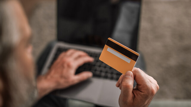 Focused Unrecognizable Senior Man Holding Credit Card While Typing On Laptop At Home Office, Engaged In Online Shopping Or Payment Process. Digital Transaction And Finance Concept.