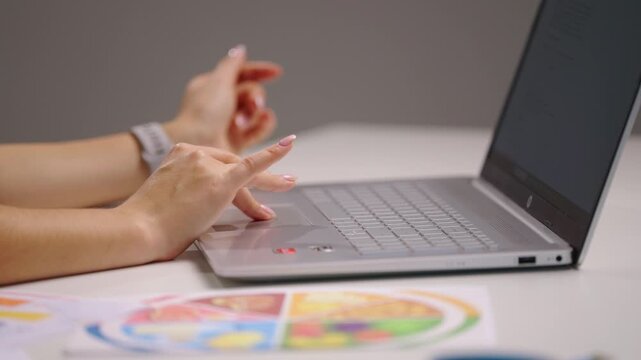 Close side view of female hands clicking and scrolling on laptop while reviewing colorful charts and diagrams, creating nutrition data analysis and planning a personalized healthy diet program