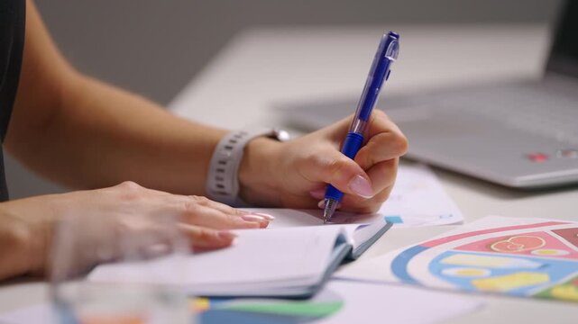 Close up of a female nutritionist writing notes in a calendar at desk with laptop and diet charts in background, planning appointments and organizing health consultation details in a modern workspace