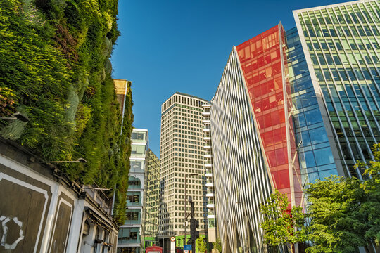 View of modern office buildings with a striking red glass facade and a lush vertical garden wall under a clear blue sky in London, England, United Kingdom.
