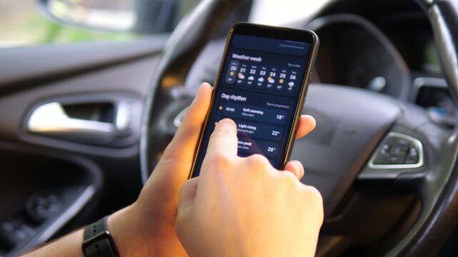 Driver scrolls through a mobile weather dashboard in a parked car, checking hourly temperature, humidity and wind trends as digital forecasts shape daily travel decisions.