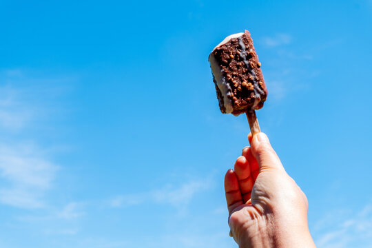 Hand holding chocolate covered ice cream bar with nuts melting on stick against bright blue sky. Cold sweet frozen dessert snack outdoors on hot sunny summer day. Ideal for refreshment design.