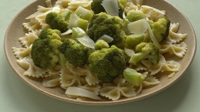 Steamed broccoli florets on farfalle pasta. Gourmet plate with garlic and parmesan on pale pastel green background, rotating smoothly in tilted view with backlit natural window light.