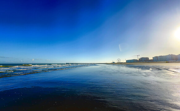 Nieuwpoort, West-Vlaanderen, Belgium, Ocotber 25th, 2025, sunrise reflection on wet sand with low sun casting warm light across shallow water, vibrant blue sky meeting shimmering surf