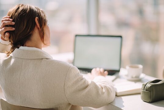 Rear view of stressed businesswoman holding her head while pausing at laptop in bright office
