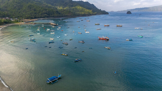 Aerial view of Pantai Karanggongso featuring numerous fishing boats anchored in the calm bay, surrounded by lush green mountains and a wooden pier in Watulimo, East Java, Indonesia.