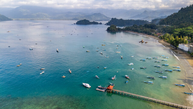 Aerial view of Pantai Karanggongso with numerous small boats anchored in the clear turquoise bay, a wooden pier extending into the water, and lush green hills in Watulimo, East Java, Indonesia.