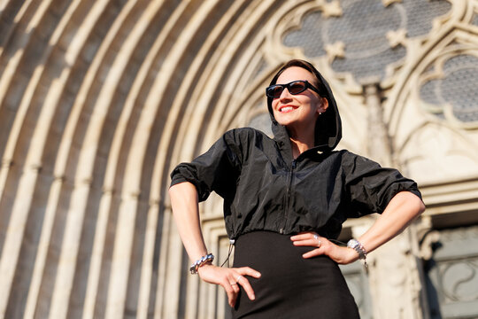 Portrait young adult elegant woman black dress and sunglasses smiles confidently in golden sunlight warm sunset backlit light against historic cathedral building. Female person walking city street