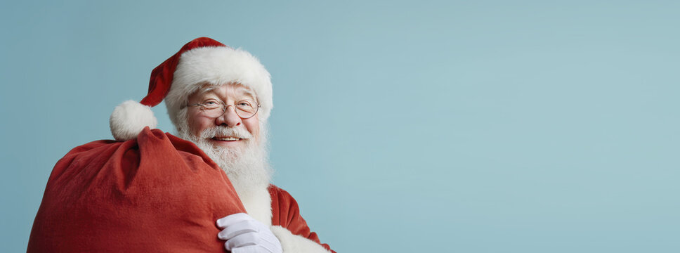 Joyful santa claus carries red sack of gifts, smiling against sky blue background
