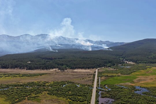 Aerial view of a wildfire burning in a dense pine forest with smoke rising against the backdrop of the Rocky Mountains, near a road and wetlands in Leadville, Colorado, United States.