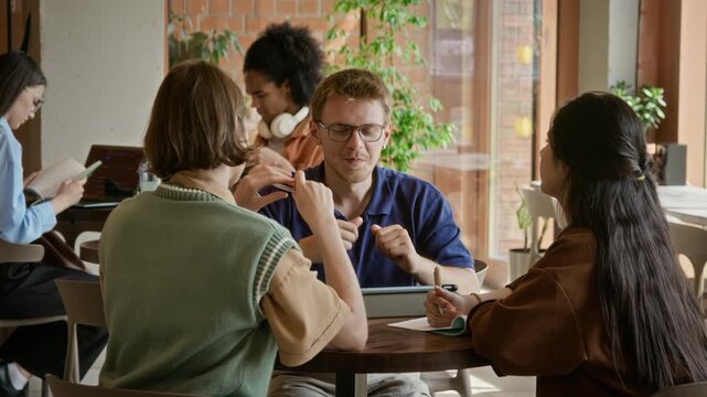 Diverse young adult students collaborating in cafe, Caucasian man explaining project to classmates using tablet. Perfect for university marketing and educational platform promotions