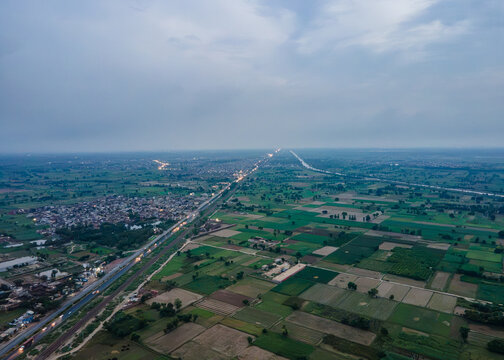 Aerial view of a rural village in Punjab, Pakistan, featuring agricultural fields, a winding road, and scattered houses under a cloudy sky.
