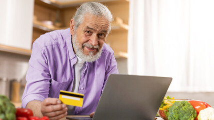 Fototapeta premium Smiling Elderly Caucasian Man Holding Credit Card While Shopping Online In Kitchen, Enjoying Modern Technology, Surrounded By Fresh Ingredients On Counter. E-commerce Concept.