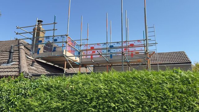 Tilt up shot against a clear blue sky, past a hedge to scaffolding poles and mesh panels, supporting a work platform of flat wooden boards, erected at house roof level, ready for work to the chimney.