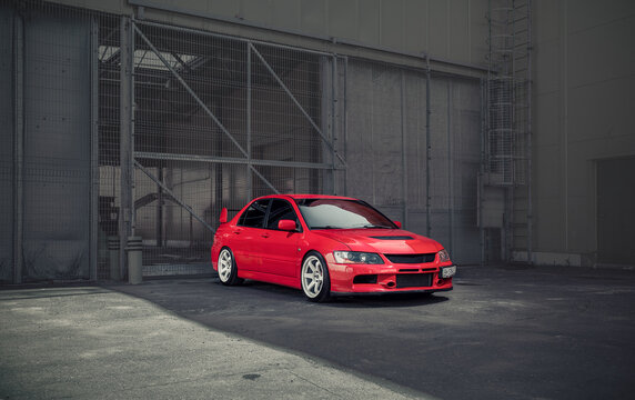 red Lancer Evolution IX parked near grey warehouse in a shadow. Three quarter front view of sporty Mitsubishi sedan in front of old industrial building gates. Odesa, Ukraine - May 07 2016