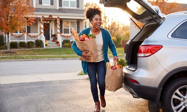 African American woman carrying grocery bags from a car at home. Thanksgiving shopping and holiday meal preparation in an autumn neighborhood. Seasonal family celebration concept
