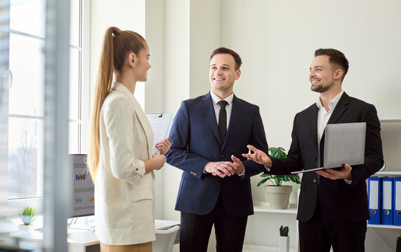 Colleagues in business office meeting share discussion. Two men welcome a woman while another presents on laptop. Bright office implies planning. Team communication concept for collaboration.