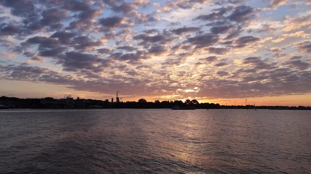 Time lapse of a spectacular summer sunrise over the IJ waterfront and port area in Amsterdam, Netherlands. Colorful morning sky with dramatic clouds reflecting warm sunlight over the water.