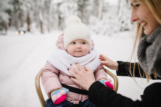 A mother adjusting a scarf on her baby, who is sitting in a sled in a snowy winter park. Close up.