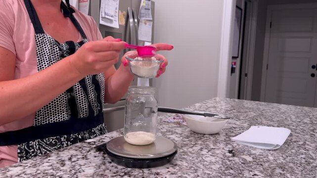 Baker weighing, feeding, discarding active sourdough starter in glass mason jars on a granite kitchen counter, preparing wild yeast for artisan baking