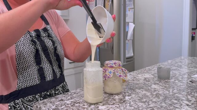 Baker weighing, feeding, discarding active sourdough starter in glass mason jars on a granite kitchen counter, preparing wild yeast for artisan baking