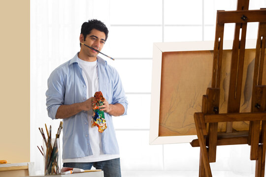Handsome young artist with paintbrush in mouth standing in front of painting 