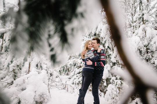 Happy couple in warm sweaters gently hugging in a snowy winter forest.