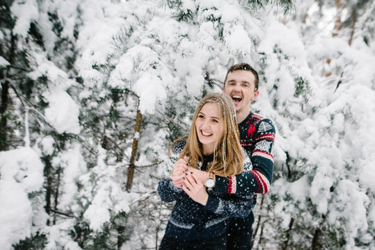 Happy loving couple, where man hugs woman from behind, they are laughing joyfully during snowfall in a snowy winter forest. Romantic moments on Valentines Day.