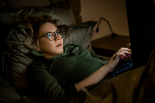 Woman in glasses typing on laptop in dark bed at night. Freelance or late work concept