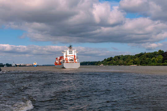 Container ship sailing along Elbe River fairway with green riverbank under cloudy sky near Port of Hamburg, global shipping and maritime trade concept