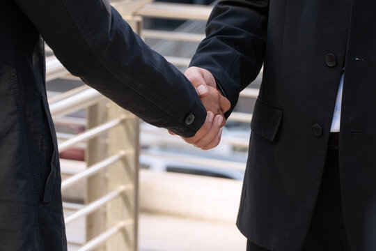 Close up of two businessmen in black suits shaking hands outdoors in the city. Corporate partnership, successful deal, and business agreement concept.
