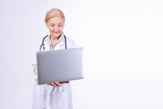 Portrait of smiling senior female doctor in white coat with stethoscope holding and using laptop computer isolated on white background copy space. Elderly healthcare professional telemedicine concept