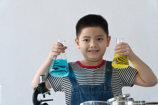Happy young Asian boy holding chemistry flasks with blue and yellow liquid. Little student doing science experiment on white background with copy space. STEM education concept.