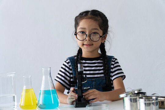 Portrait of a smart little Asian girl wearing glasses doing science experiment with microscope and colorful liquid flasks on desk isolated on white background. STEM education concept.
