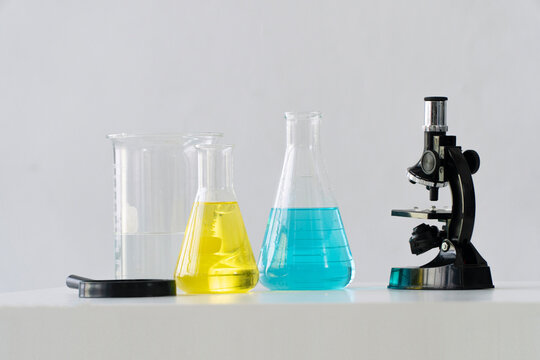 Science laboratory equipment including chemistry flasks blue, yellow liquid, clear beaker, magnifying glass, microscope on table isolated on white background.STEM education scientific research concept