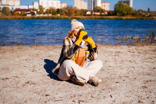 Woman in warm clothes drinks hot tea from thermos cup sitting on sandy beach. Sunny autumn day outdoors by the water.