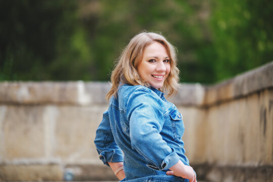 Smiling woman in denim jacket stands near stone bridge turning back smiling at camera. Concept of confident urban lifestyle and positive mindset.