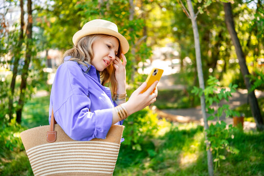 Adult woman in straw hat holds striped bag looking at smartphone screen with squinting eyes. Concept of bad eyesight and digital device problems.