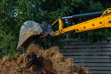 Close up of yellow excavator bucket dumping dirt soil on pile near backyard wooden board fence. Residential excavation works  © Marya