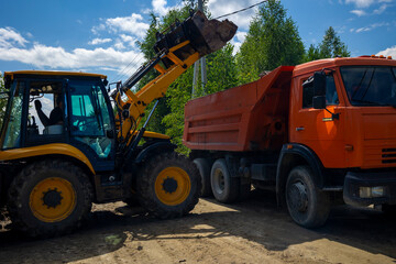 Yellow backhoe loader filling dumper truck with dirt soil for construction site clearing. Concept: site clearing operation © Marya