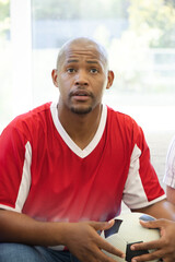 Fototapeta premium African American man holding black-white soccer ball on sofa in red white jersey, looking at window