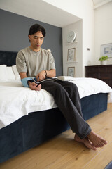 Fototapeta premium Asian man sitting on bed edge in bedroom adjusting light-blue blood pressure cuff and white monitor