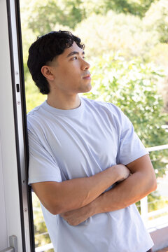 Asian man leaning against door jamb wearing light blue T-shirt, gazing toward balcony railing