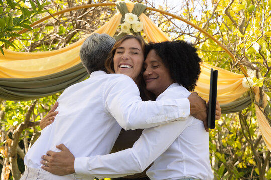 Diverse group hugging under arch draped yellow olive fabric white flowers in park right with tablet