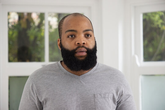 African American man standing by white-framed windows at home, wearing gray shirt with chest pocket