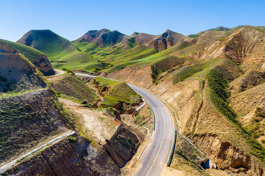 Winding asphalt road through eroded green hills and dry ravines in Xinjiang, China, offering travel, infrastructure, and rural landscape appeal.