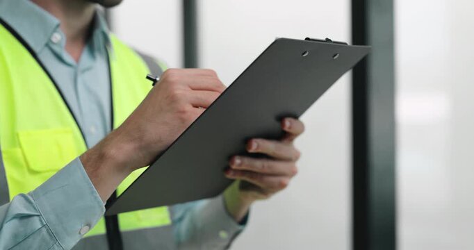 Construction worker writes inspection notes on clipboard in bright workspace. Engineer holds board firmly as records project details with careful focus