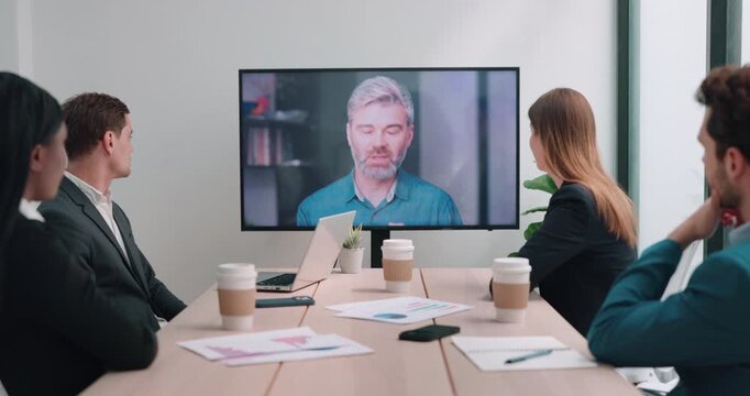 Diverse business team listens to remote Caucasian boss on screen during video conference. Colleagues watch manager speaking continuing focused online meeting