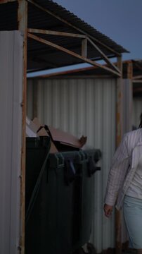 Young female casually discards disposable cup into overflowing waste bin filled with boxes during evening near corrugated shelter in urban setting.